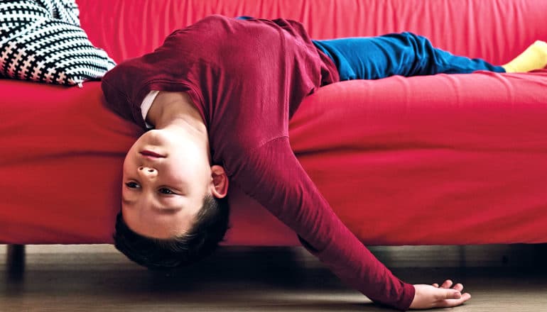 A boy lays upside down on a red couch looking bored