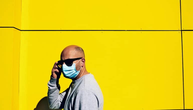 A man in a face mask and sunglasses walks past a bright yellow wall