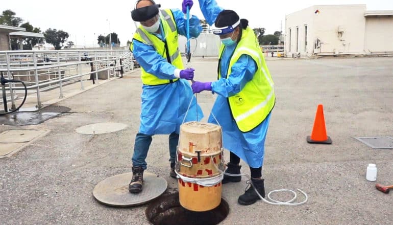 Two workers in protective gear lower a container into a sewer entrance with a rope