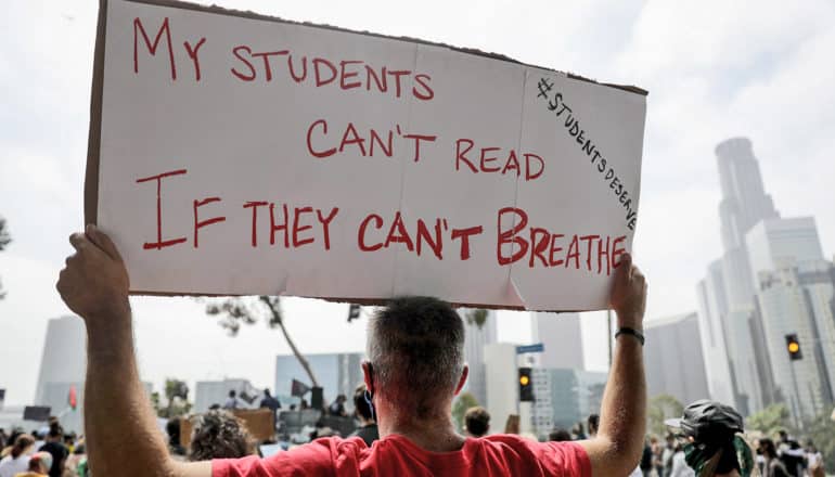 person holds sign reading "my students can't read if they can't breath #StudentsDeserve"