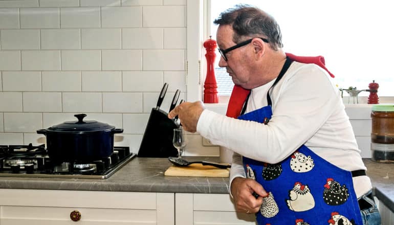 older man wearing apron points to pot on stove