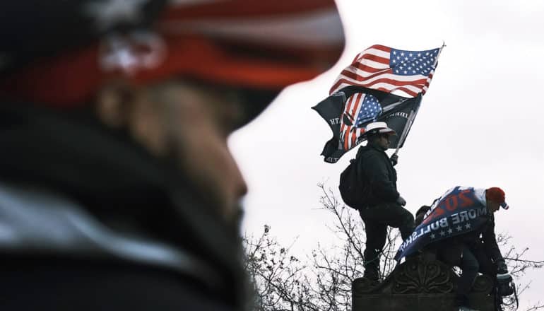 blurry person in US flag hat in foreground; background shows two people on statue with US flag and "Punisher" flag