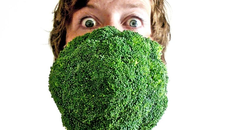 A woman has a giant broccoli floret in her mouth and a surprised look in her eyes