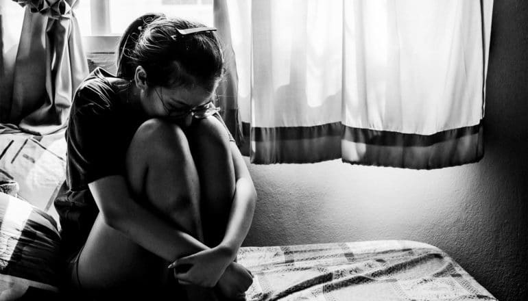 A young teen girl sits on her bed with her head on her knees