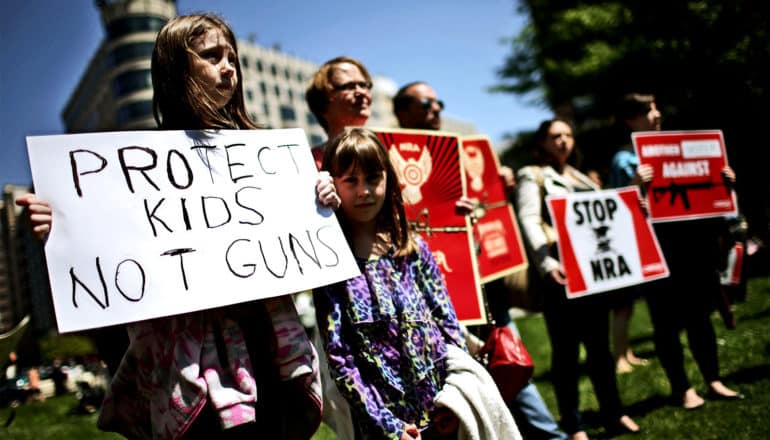 Two young girls hold a sign that reads "Protect kids not guns" while standing with other gun control protestors