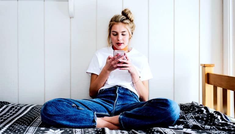 A young woman reads her phone while sitting on her bed