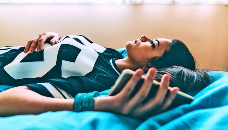 A young woman lays on her bed holding her phone staring into space