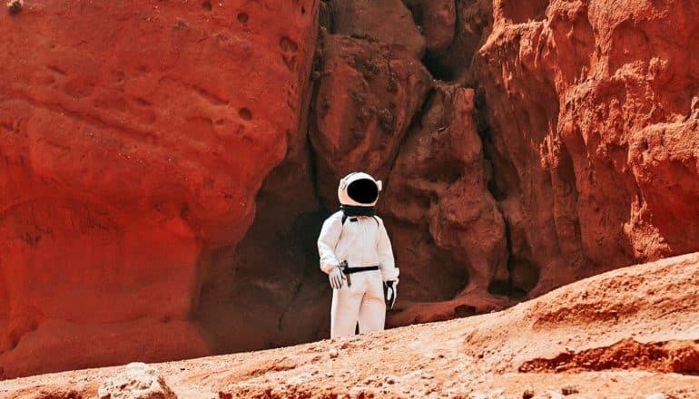 A lone astronaut walks through a red, rocky environment that looks like Mars