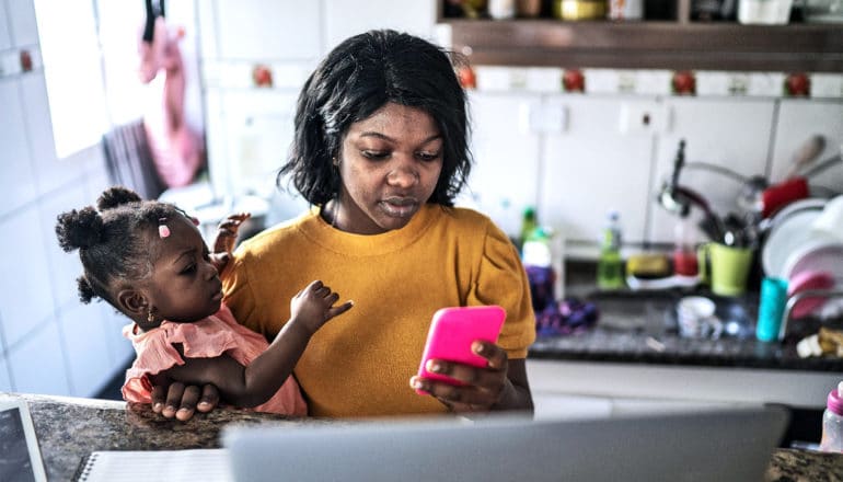 A young woman holds her baby while working from home in her kitchen