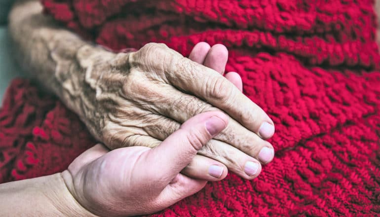 An older cancer patient holds the hand of a family member against a red blanket