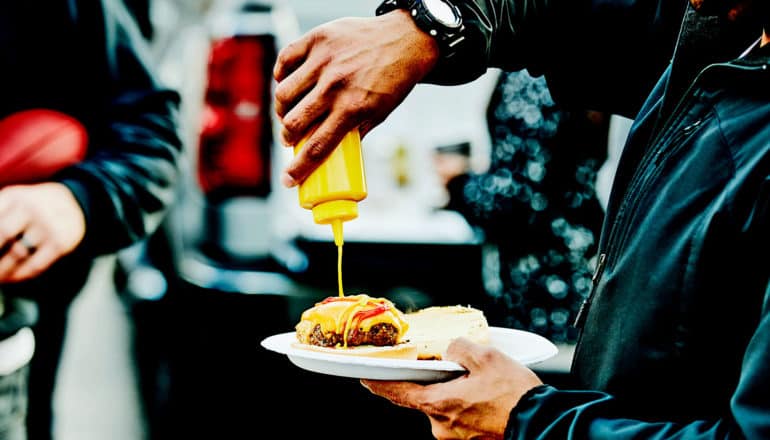 A man squeezes mustard onto a burger while tailgating