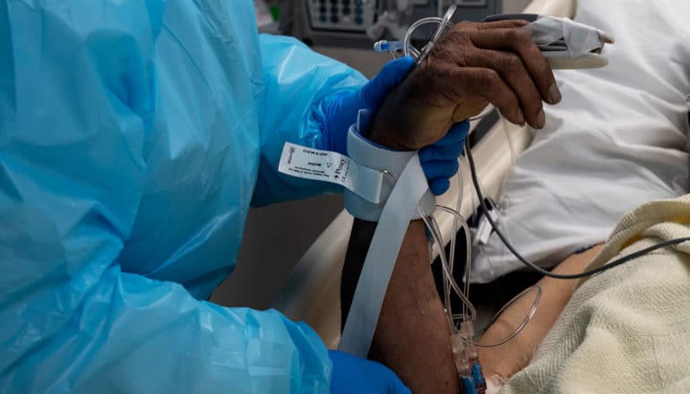 health care workers in blue PPE holds up hand of COVID patient in hospital bed