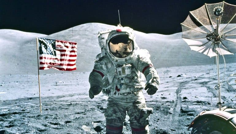 An astronaut stands on the surface of the moon next to an American flag and a rover