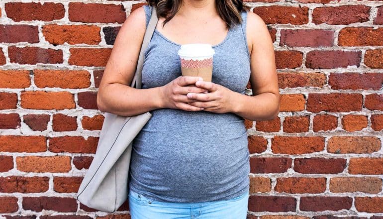 A pregnant person holds a coffee cup while standing against a brick wall