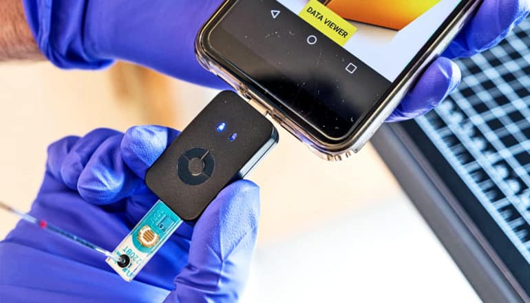 A researcher wearing purple gloves holds the small black device plugged into the phone. The device has a blue chip coming out of the bottom.