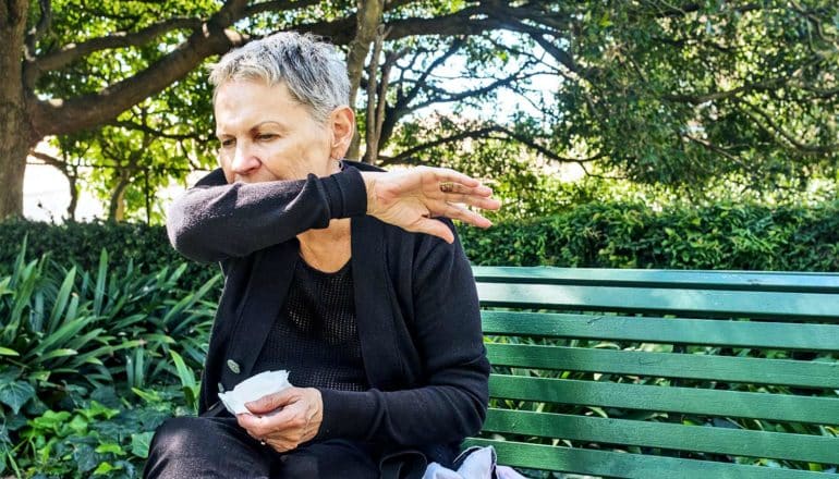 An older woman on a park bench coughs into her arm