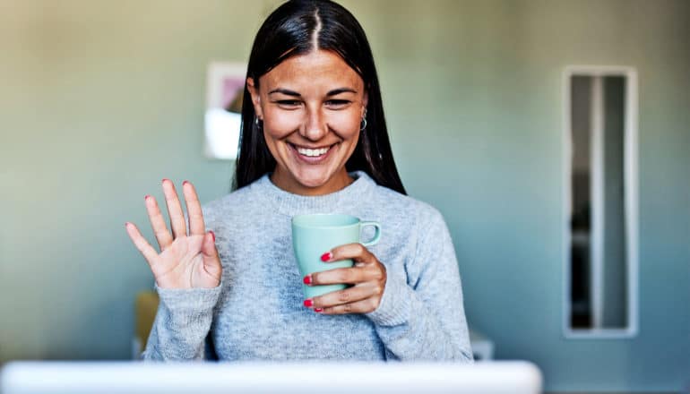 A young woman holds a coffee cup and waves to her laptop during a virtual meeting