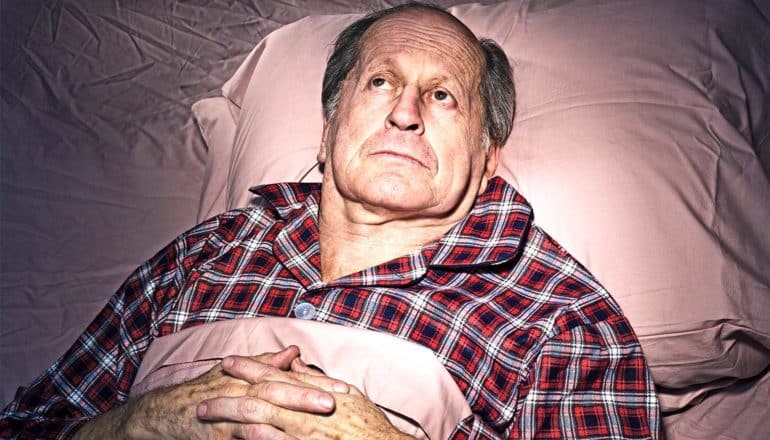 A man lays in bed with his eyes open and hands folded