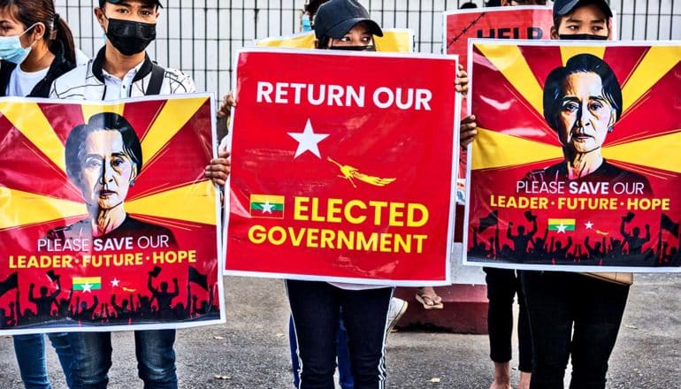 Protestors hold signs that call for the restoration of Myanmar's civilian government, one reading "Return Our Elected Government" and the others with pictures of Syu Kyi