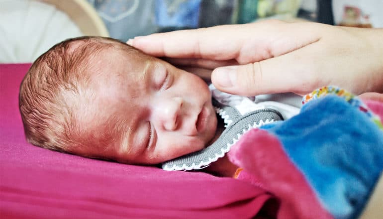 A mother touches her preterm baby's cheek while the baby lays on a pink blanket