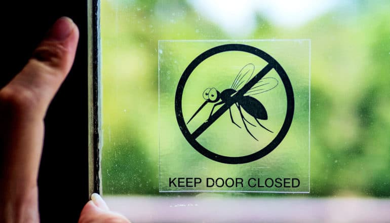 A woman closes a door with a sticker on it that has a mosquito on it and reads "Keep Door Closed"