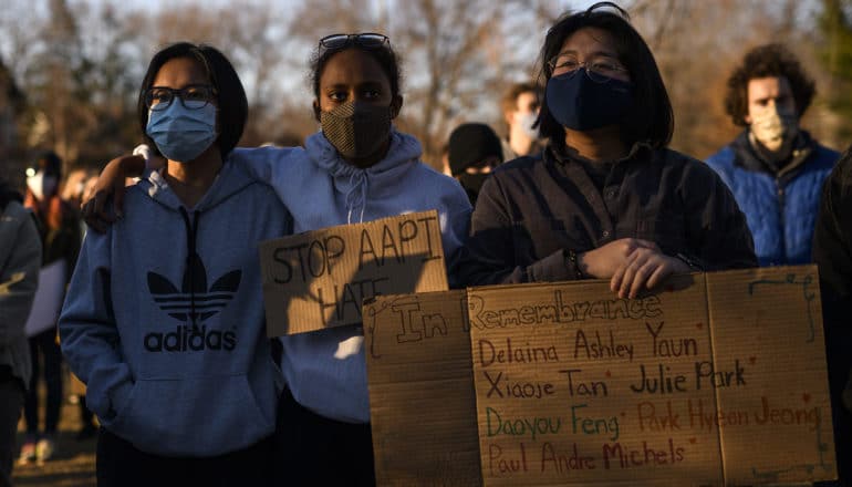 three masked people stand solemly, one with sign with names of victims of Atlanta spa shooting