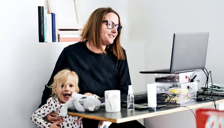 A woman in a home office looks at her laptop screen while holding her daughter