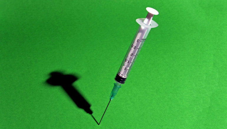 A syringe sticking into a green surface with its shadow in the background