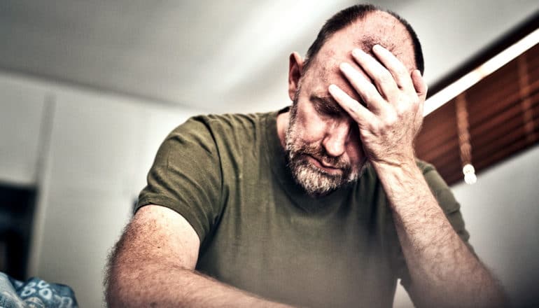 A man holds his forehead while sitting on his bed