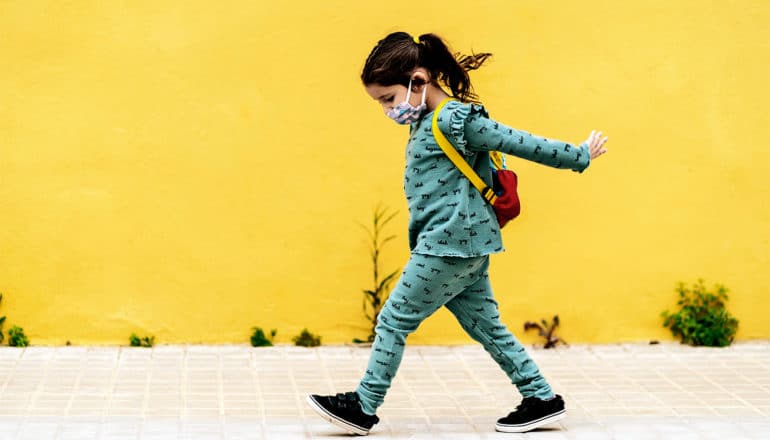 A little girl wearing a face mask and backpack walks past a yellow wall