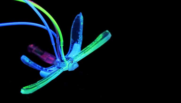 The soft robot is lighted in different colors, green, blue, and purple. It has two sets of wings and a long tail, with thin wires coming from its body