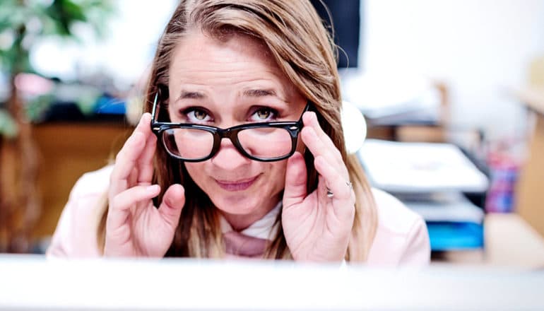 A woman looking up from a computer screen touches her glasses and looks skeptical