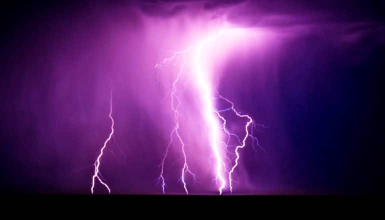 Lightning flashes across a dark sky, making clouds appear purple