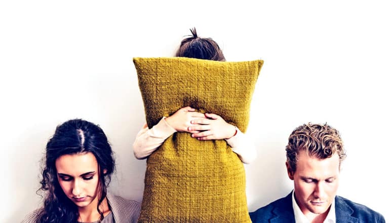 A young girl holds a pillow over her face while her parents sit on either side of her looking down