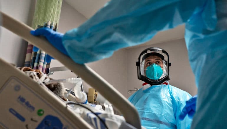 health care provider in PPE across patient bed. Arm of another caregiver in foreground