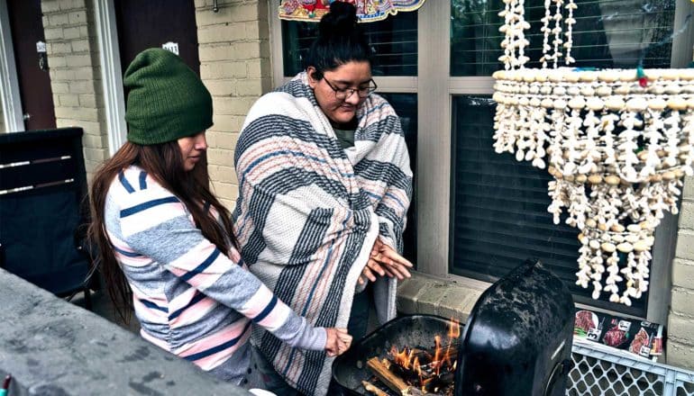 Two people warm their hands on a wood-fire grill on their porch