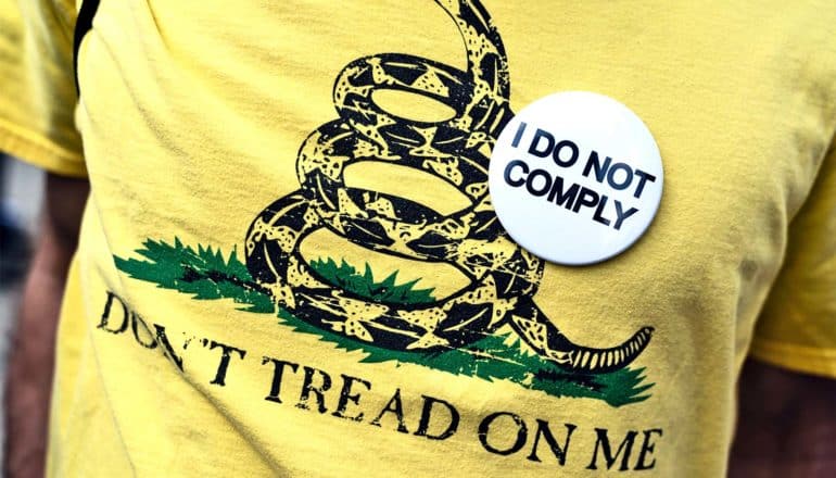 A man wears a yellow "Don't Tread On Me" t-shirt and a pin that reads "I do not comply"