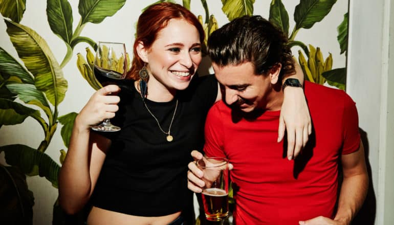 grinning young woman holds wine glass and drapes arm around neck of young man holding beer