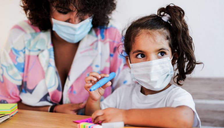 adult and young child in masks sit at table with markers