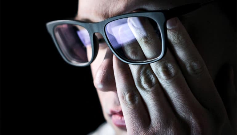 A man rubs his left eye as his glasses reflect a computer screen