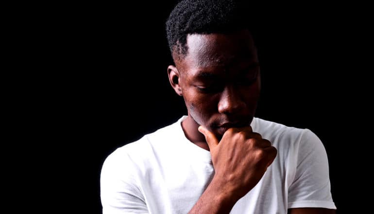 A young Black man half in shadow looks down with his hand on his chin