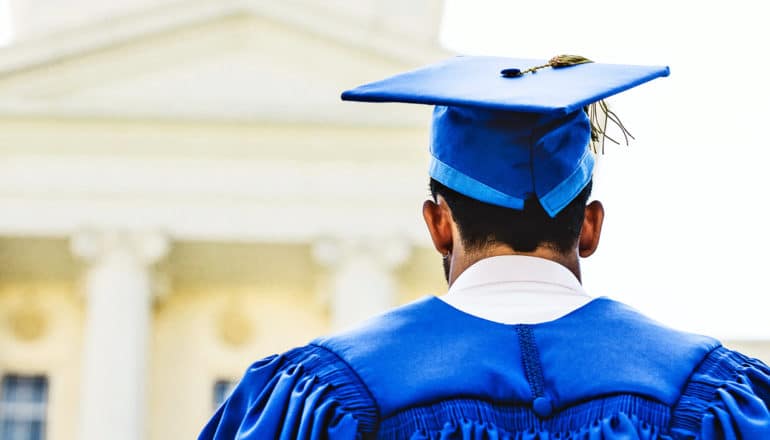 A young man in a blue graduation cap and gown looks up at a university building