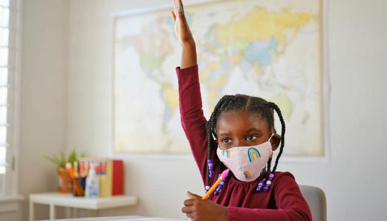 Black child in rainbow mask raises hand in classroom