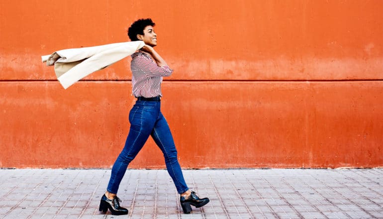 A woman walks past an orange wall tossing her coat over her shoulder