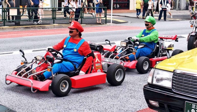 Two people dressed as Mario Kart characters drive go carts through a street