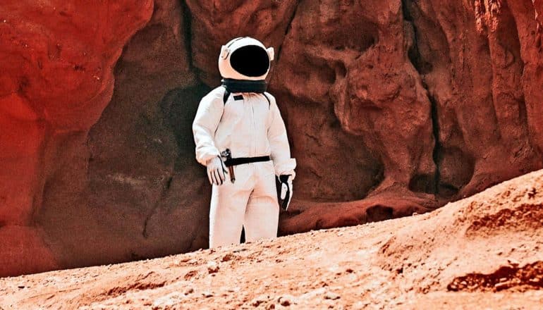 A person in an astronaut costume walks across red dirt with red rocks in the background