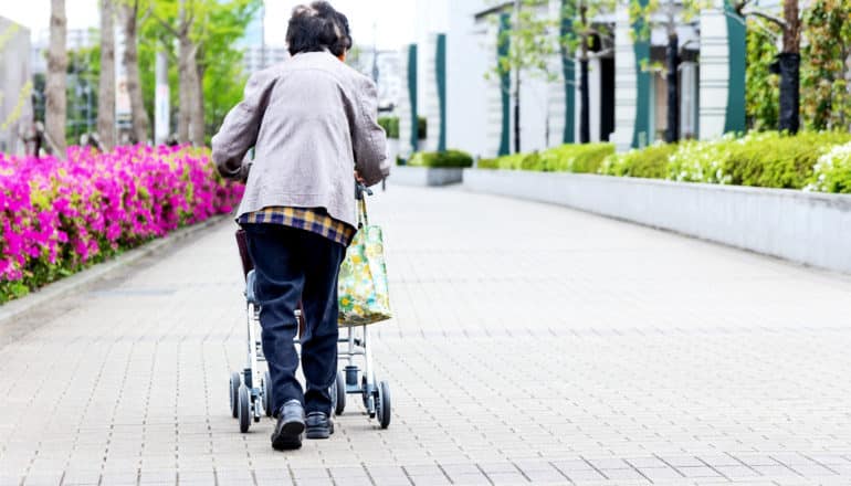 An older woman walks down a sidewalk with a rolling walker