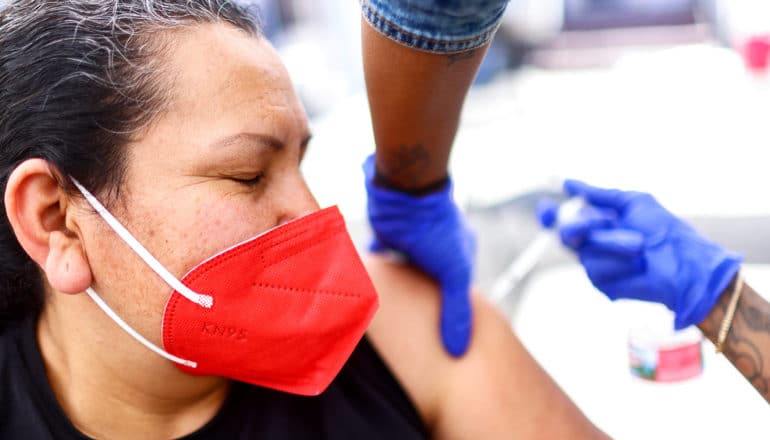 person in red KN95 mask closes eyes as blue-gloved hands inject vaccine into arm