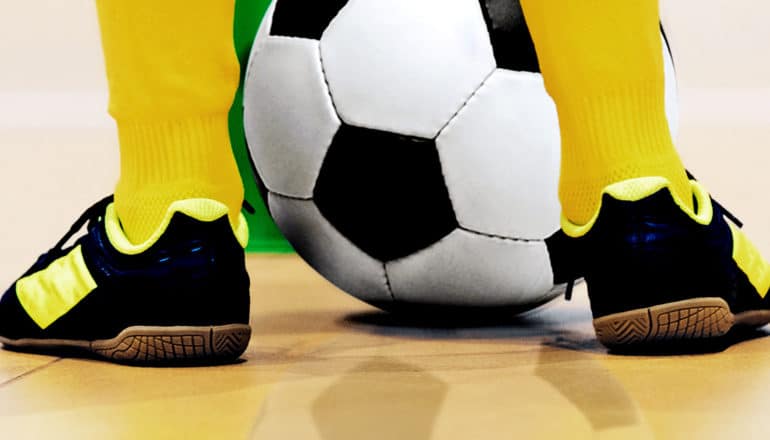 A young student stands in a gym with a soccer ball between his feet