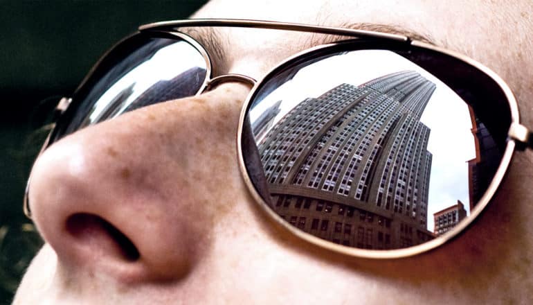 A woman wearing glasses looks up at a building, visible in the reflection of the glasses